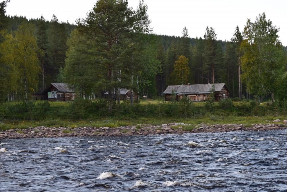 In the foreground, a river flows, and on the other bank, there are wooden red buildings.