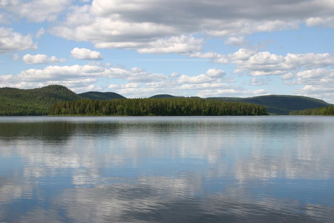 Tyyni järvi, jonka pintaan piirtyy hennosti taivaalla olevat pilvet. Takana kohoaa metsäisiä mäkiä.