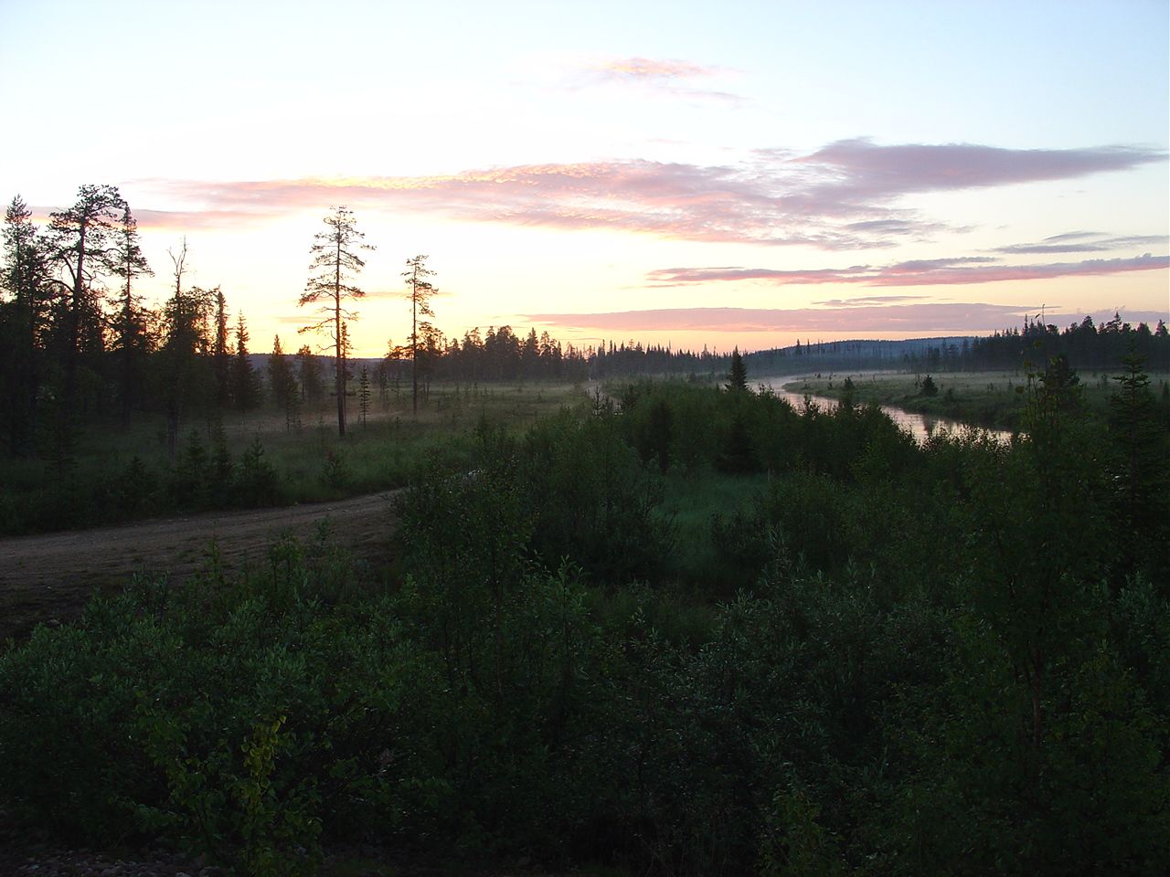 Maisema iltahämärässä. Suon ja kapean joen välissä kulkee metsäautotie. Aurinko laskee horisonttiin.