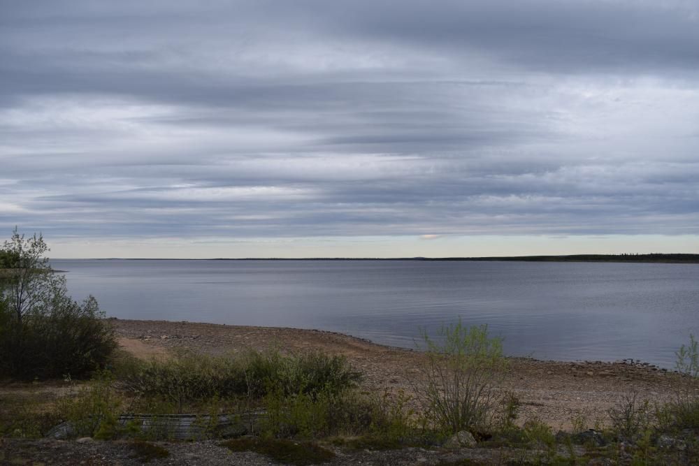 Kivisen rannan takana aukeaa tyyni iso järvi. Rannalla kasvaa matalia puskia ja muuta kasvillisuutta.