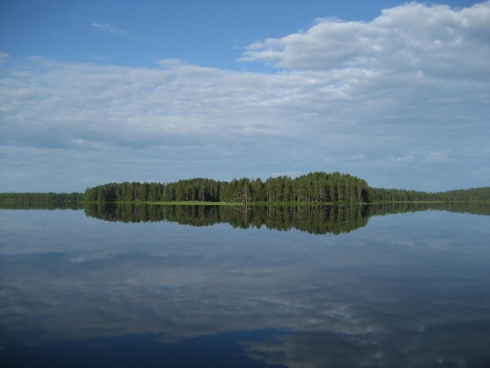 Peilityyni järven pinta, johon heijastuu taivaalla olevat pilvet ja vastarannan metsä.