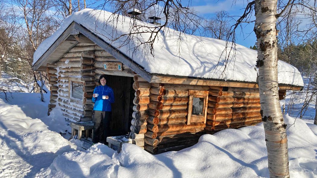 A log cabin with snow on the roof. A woman is standing in front of the cabin.