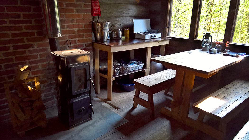 Inside the building there is a table, benches and a kitchenette. From the windows one can see summer forest.