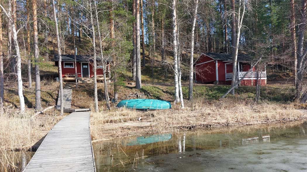Lammasharjun pihapiiri laiturilta nähtynä. Pihassa on mökki ja sauna, rannassa soutuvene.