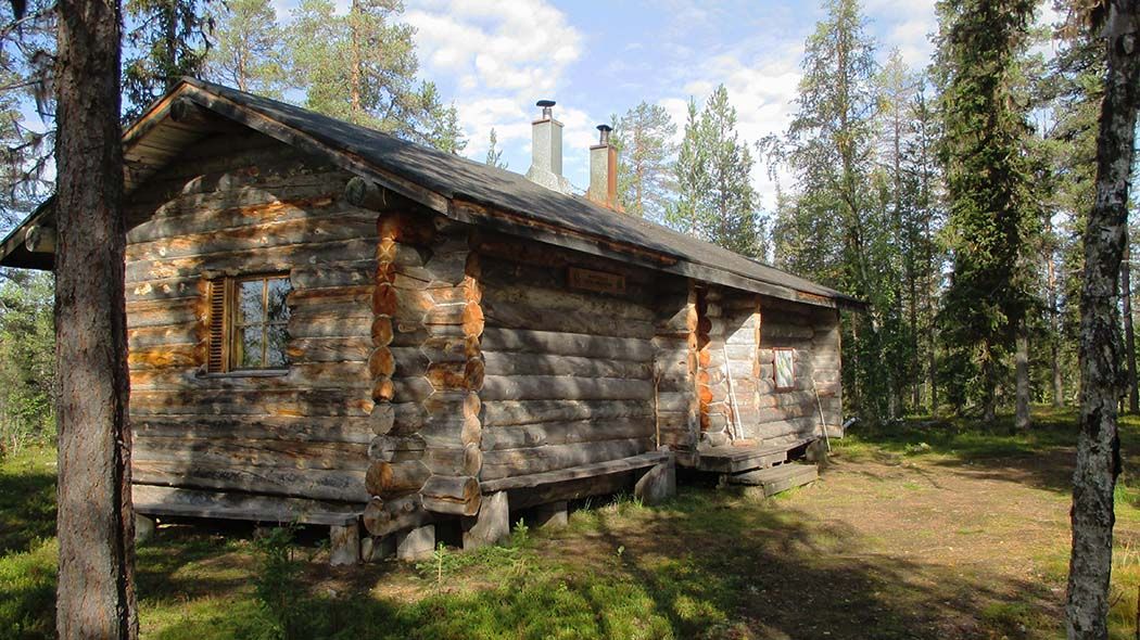 Hirsisen kämpän seinustalla on penkit. Päätyseinällä on ikkuna, jossa ei ole verhoja. Tuvan katolta nousee kaksi savupiippua.
