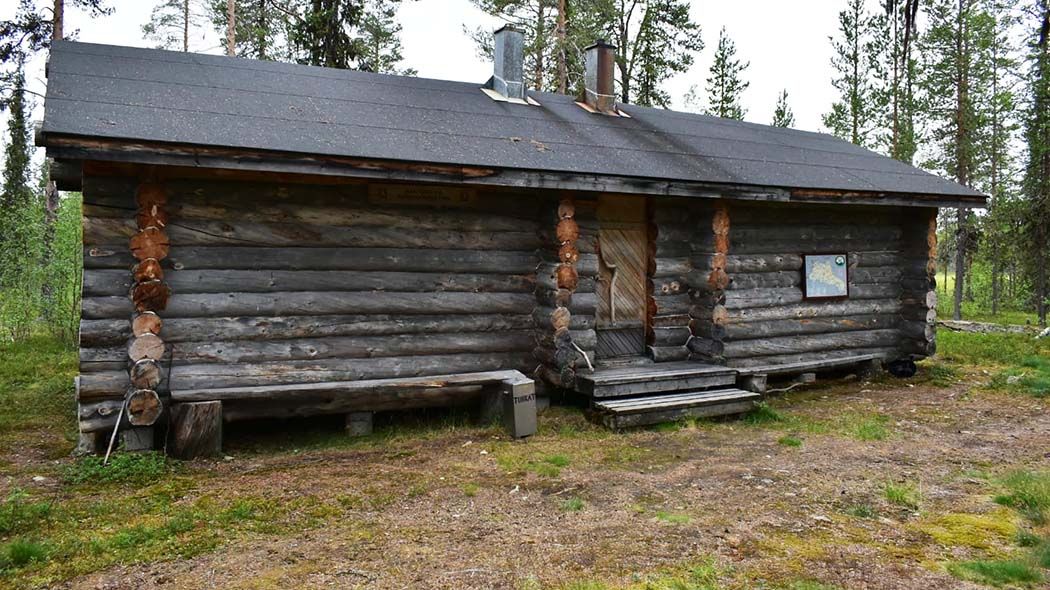 Hirsinen kämppä, jossa on ovi. Oven oikealla puolella on taulu seinällä ja vasemmalla puolella maassa peltinen kannellinen astia.