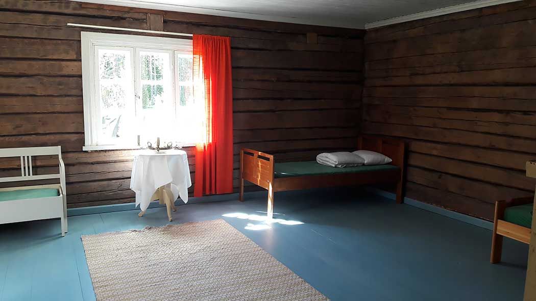 Inside in front of the hall window, there is a small table, a bed in the corner and a rug on the floor.
