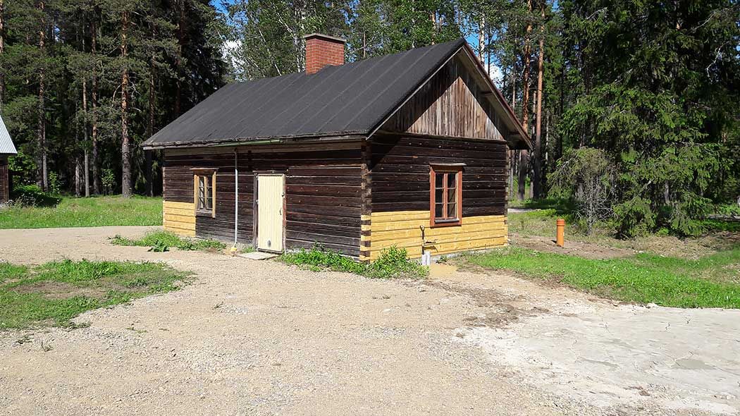 A wooden building with a chimney on the roof in summer weather. A forest in the background.