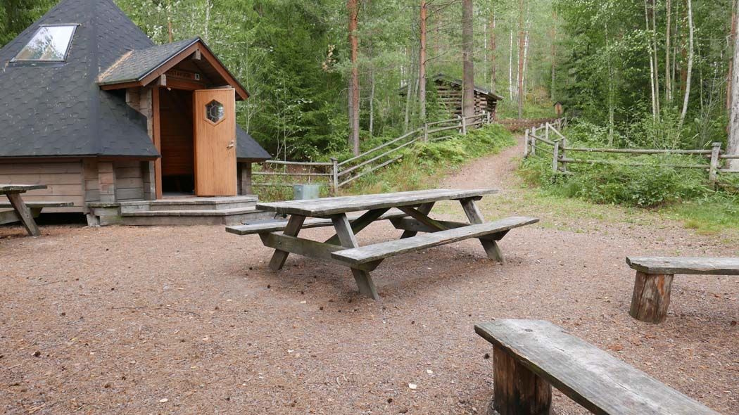 Aidatun pihan laidalla on kuusikulmainen hirsikota. Ovi on avoinna.