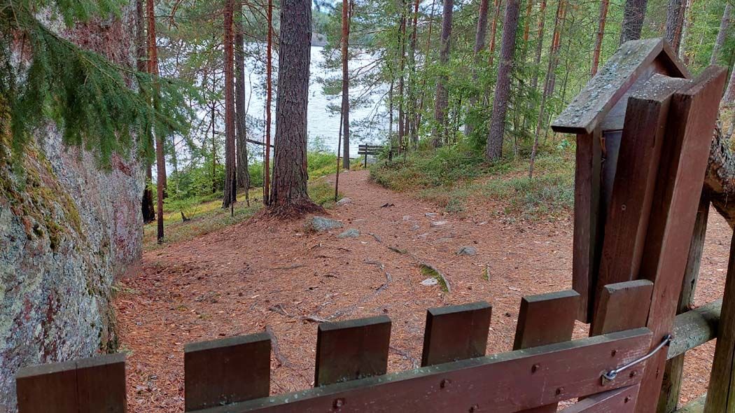 Kuvan etualalla on ruskea portti. Portin takana kapea metsäpolku johtaa mäkeä alas rantaan. Rannassa häämöttää puinen laituri.