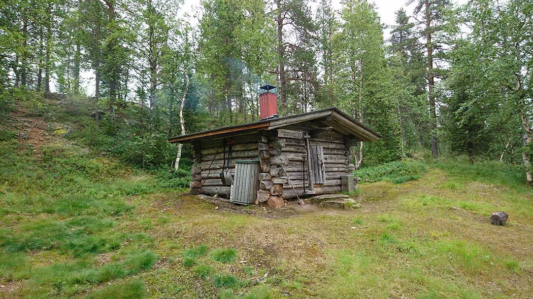 Rök stiger upp ur skorstenen i en liten timmerstuga. Det finns en liten dörr i mitten av huvudväggen. Bakom huset finns en skogsklädd sluttning.