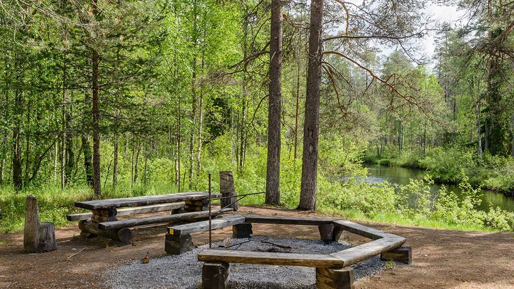 En eldplats vid stranden. Bredvid står ett bord med bänkar. I bakgrunden finns skog.