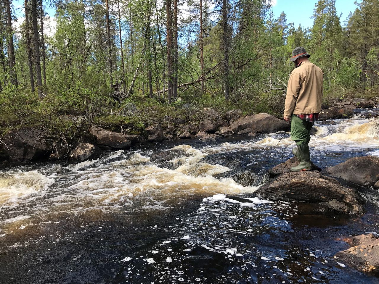 En man står på en stor sten i floden. Floden strömmar snabbt.