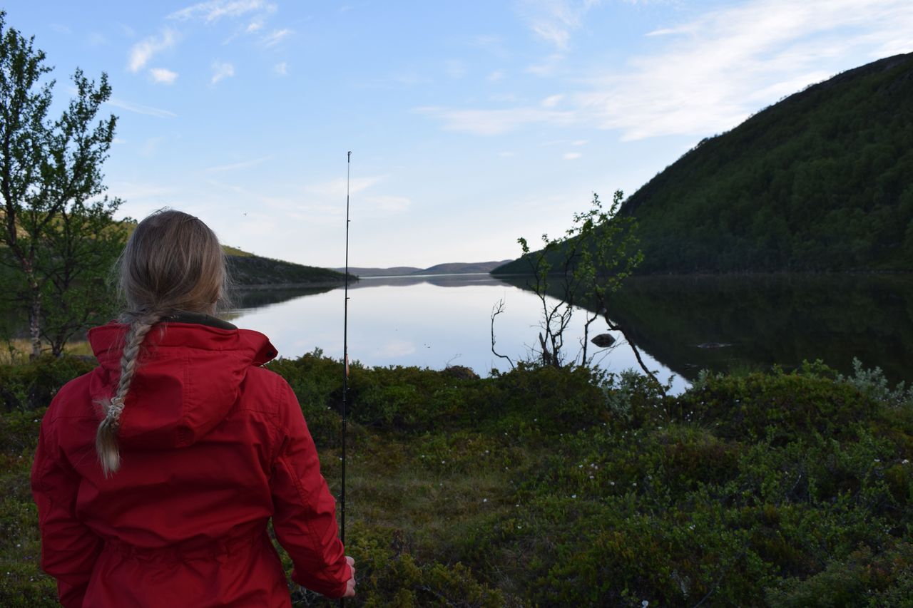 A fisher stands with her back to the camera, holding a rod and wearing a red jacket. Behind her, a lake and a fell landscape open up. The sky is blue and clear.