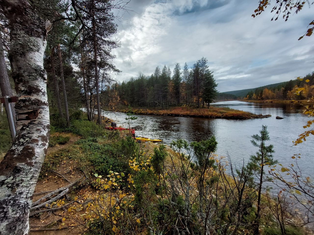 Syksyinen jokimaisema. Maassa ja puissa on ruskan värejä, taivas on pilvinen.
