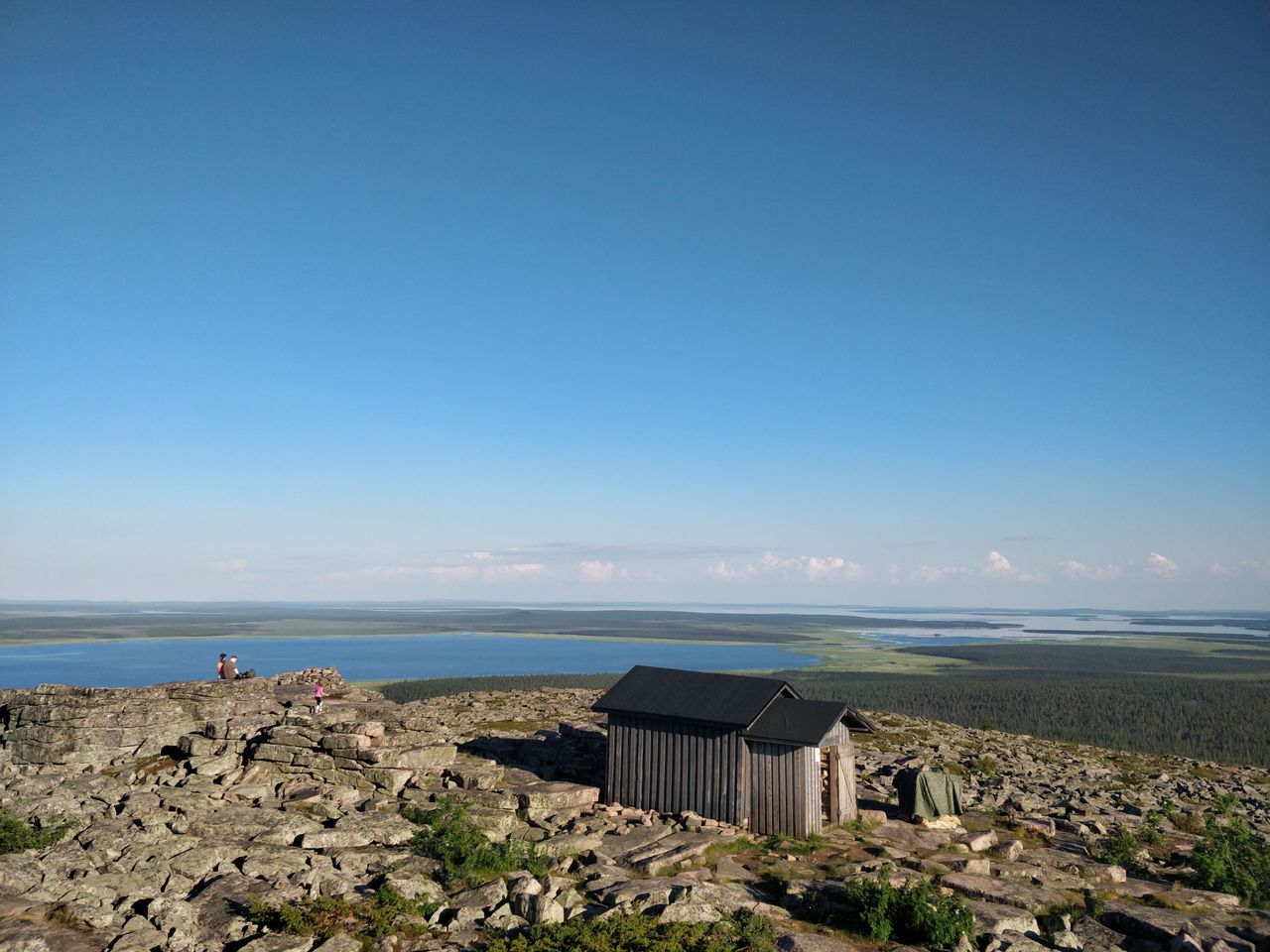 Tunturin kivinen huippu ja autiotupa aurinkoisena kesäpäivänä. Taaempana alhaalla näkyy iso järvi.