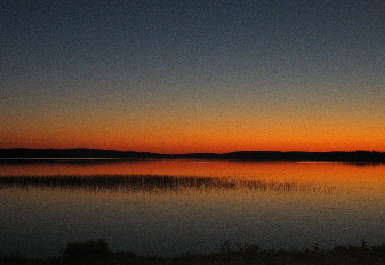 Auringonlasku on värittänyt taivaan ja tyynen järven pinnan oranssiksi.