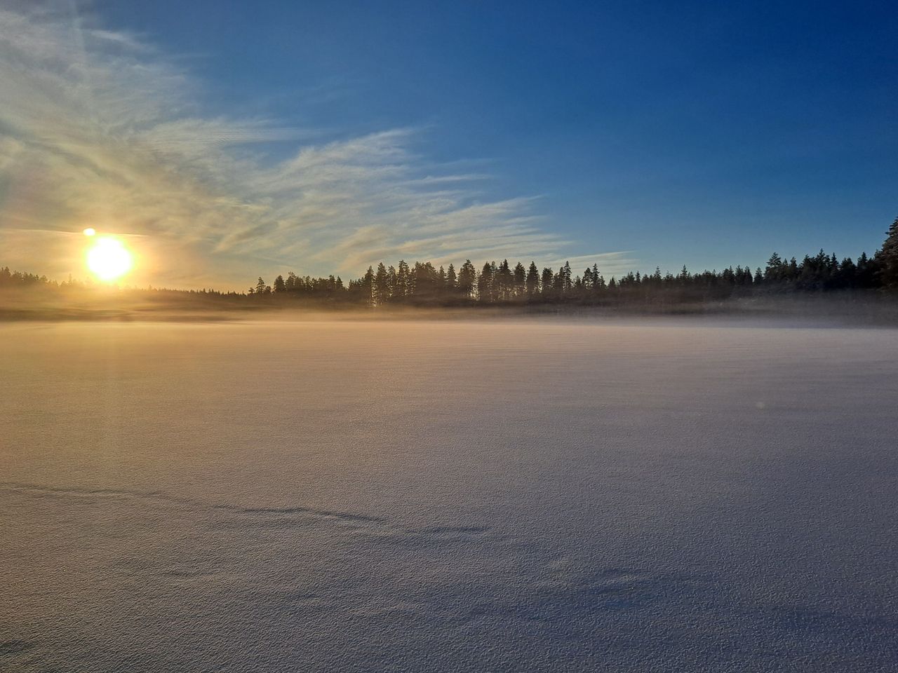 The lake is covered with snow and ice. In the distance, there are forests growing on the shore. The sun is shining and the sky is blue.