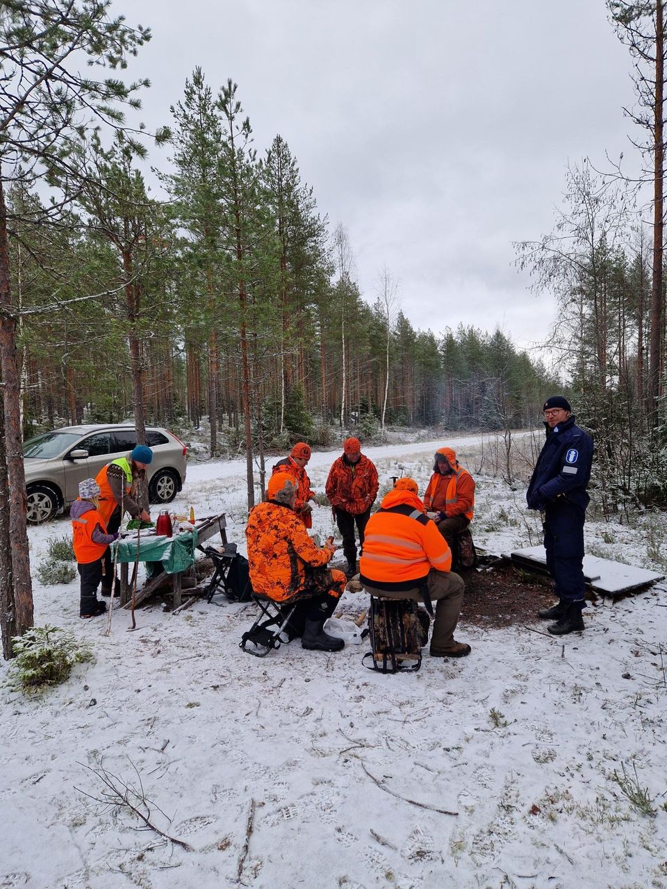 Five hunters in orange outfits are sitting by the campfire on a break. Behind them, an adult and a child are preparing snacks at a table. Next to the hunters stands a police officer who is smiling and looking at the camera.