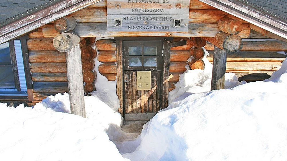 Hirsituvan sisäänkäynti lähikuvassa. Tupa on lumikinosten ympäröimä ja ovelle johtaa vain kapea polku. Oven yläpuolella on katos, johon on kiinnitetty kyltit, joissa lukee: "Metsähallitus Pöyrisjärvi" ja "Meahceráđđehus Bievrrašjávrri".