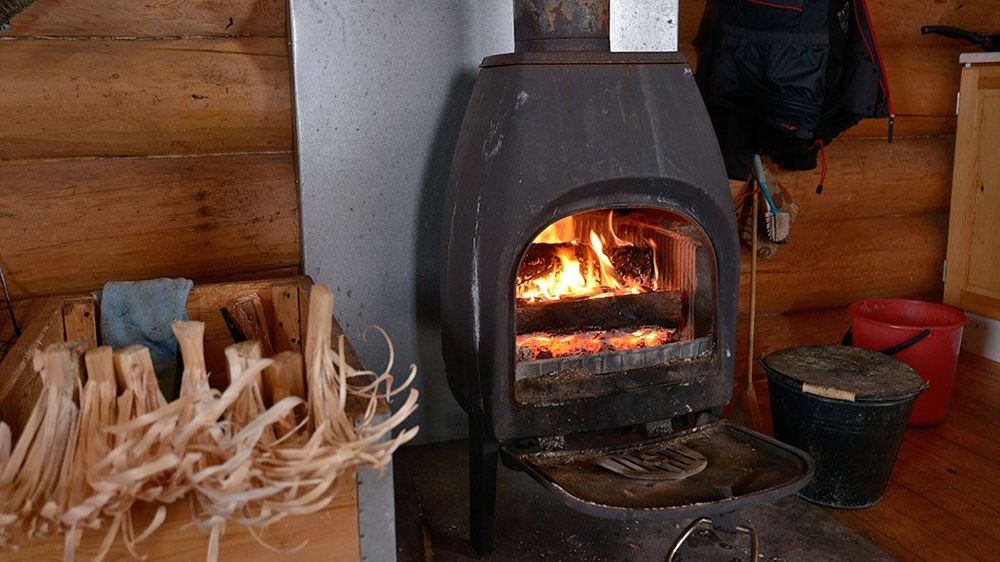 Tuvan kaminassa loimuaa tuli. Kaminassa on suuri luukku, joka on avoinna. Etualalla on valmiiksi tehtyjä kiehisiä.
