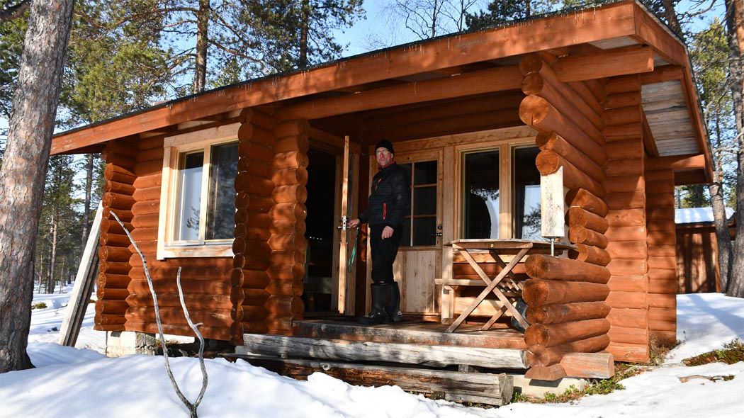 Det står en person på trappan till bastubyggnaden i rundtimmer och öppnar dörren. Solen skiner och det ligger snö på marken. Stora tallar växer runt byggnaden.